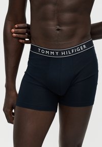 Ανδρικό μοντέλο φοράει σκούρα μπλε boxer Tommy Hilfiger με λογότυπο στη μέση, στέκεται μπροστά σε απλό ανοιχτό φόντο.