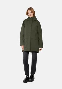 Olivgrüner, wasserdichter Mantel mit Kapuze, zwei vorderen Taschen und geradem Schnitt. Getragen über dunklen Hosen und schwarzen Stiefeletten.