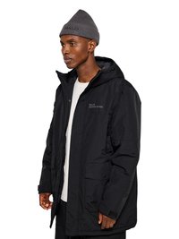 Mies, jolla on harmaa pipo ja musta hupullinen talvitakki edessä taskuilla sekä valkoinen paita, katsoo vasemmalle valkoista taustaa vasten.