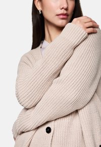 Hellbeige gestrickte Strickjacke mit strukturiertem Muster, langen Ärmeln und einer vorderen Knopfleiste. Weicher, gemütlicher Stoff mit gerippten Akzenten.