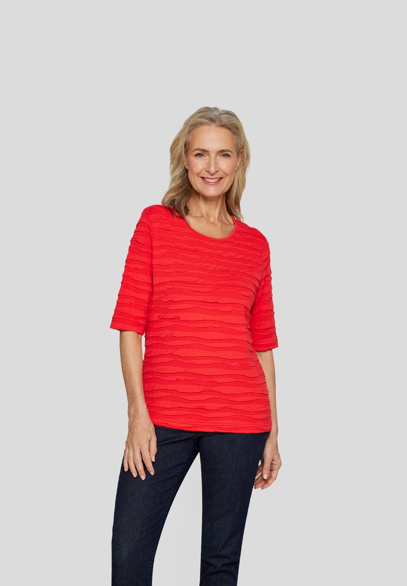 Rotes oberteil mit strukturiertem Muster und wellenförmigen Streifen, runder Ausschnitt und Dreiviertelärmel, kombiniert mit dunklen Jeans. Schlichtes Design und lebendige Farbe.