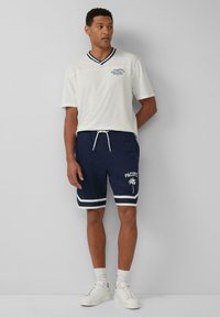 Wit shirt met korte mouwen en een marineblauwe V-hals, gecombineerd met marineblauwe shorts met witte accenten en de tekst "PACIFIC" met een palmbomen ontwerp. Witte sneakers.