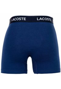 Granatowe bawełniane szorty typu bokserki z czarną gumką w talii, na której znajduje się białe logo "LACOSTE". Gładka faktura i dopasowany krój.