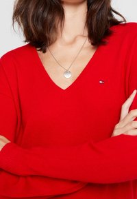 Pull en tricot rouge avec un col en V et des manches longues. Pr�ésente une petite patch logo sur la poitrine et des poignets de manches texturés. Collier en argent visible.