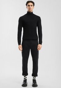 Man staat in een zwarte coltrui, zwarte opgerolde broek, zwarte schoenen en zwart-wit geblokte sokken tegen een effen lichte achtergrond.