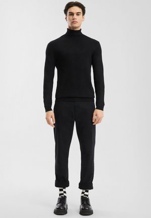 Mann steht in schwarzem Rollkragenpullover, schwarzen hochgekrempelten Hosen, schwarzen Schuhen und schwarz-weiß karierten Socken vor einem schlichten hellen Hintergrund.