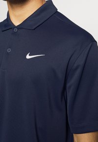 Polo bleu marine à texture douce, avec trois boutons au col et un logo Nike blanc sur la poitrine gauche. Manches courtes et coupe ajustée.