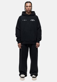 Zwarte hoodie met voorzak en logo's. Gecombineerd met loszittende zwarte joggingsbroek en casual schoenen. Zachte stofstructuur.