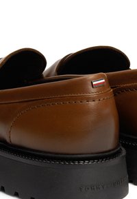 Braune Lederschuhe mit runder Form, schwarzer Gummisohle und einem kleinen Flaggenakzent an der Ferse. Sichtbare Steppnähte entlang der Nähte.