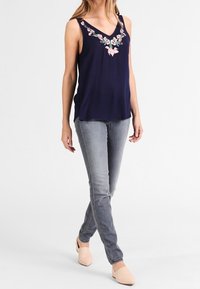 A woman walks confidently, wearing a navy blouse with floral embroidery, gray jeans, and light-colored flats, against a plain background.