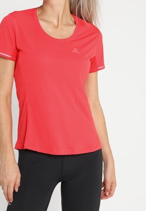 Kvinde iført en tætsiddende rød sportst-shirt med korte ærmer og sorte leggings, stående mod en ensfarvet lys baggrund.