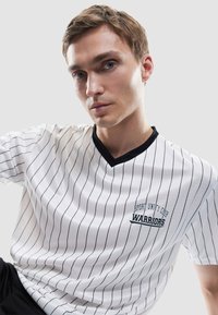 Joven con pelo corto castaño que lleva una camiseta blanca de cuello en V con rayas verticales negras y el texto "Sport Unity Club Warriors".