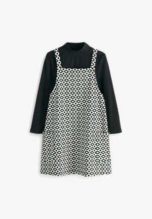 Enfant portant un haut à manches longues noir, des leggings noirs, une robe tablier fleurie noire et blanche, et des bottines à lacets rouge foncé brillant, en train de marcher.