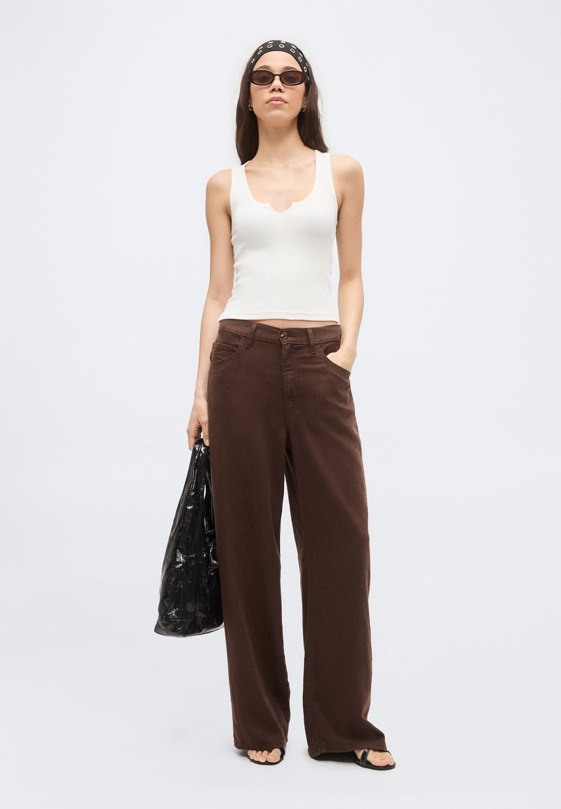 Woman wearing white sleeveless top, brown wide-leg pants, black sunglasses, and headband, holding a black tote bag, standing on plain background.