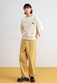 Sweatshirt crème avec un petit patch brun, associé à un pantalon jaune ample et des chaussures noires épaisses, sur un fond uni.