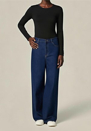 Personne à la peau foncée portant un haut noir à manches longues, un jean bleu taille haute à jambes larges, et des chaussures blanches, debout avec une main sur la hanche.