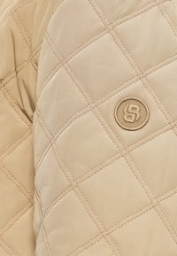 Béžová prešívaná bunda s diamantovým vzorom a hnedým prešitím, s kruhovým vyšívaným logom na látke.