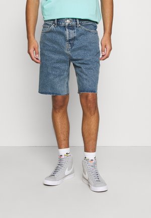 Shorts en denim bleu clair avec un ourlet effiloché, portés avec des baskets montantes grises et des chaussettes blanches avec un logo. Design simple à cinq poches.