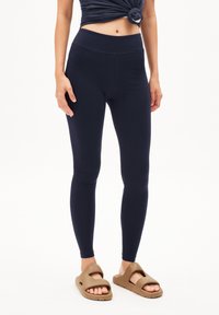 Marineblauwe legging met een hoge tailleband, gemaakt van een gladde, rekbare stof. Draag ze met bruine instapsandalen.