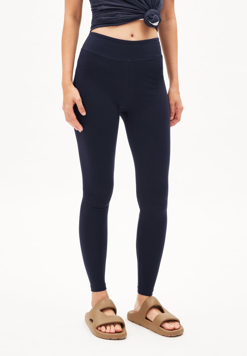 Marineblauwe legging met een hoge tailleband, gemaakt van een gladde, rekbare stof. Draag ze met bruine instapsandalen.