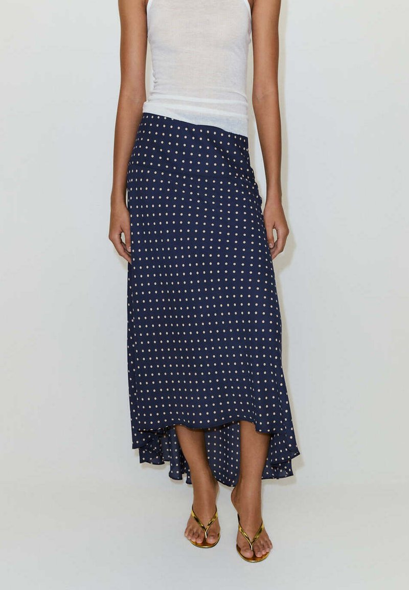 Femme portant une jupe bleu marine à pois style 'high-low', associée à un top blanc sans manches et des sandales dorées, debout devant un fond uni.