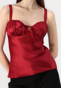 Camisola de satén rojo con escote corazón acanalado, tirantes ajustables y un detalle de lazo en el centro. Textura suave, silueta ajustada.
