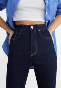 Dark blue denim high-waisted jeans with orange stitching and a front button closure. Features side pockets and a fitted silhouette.