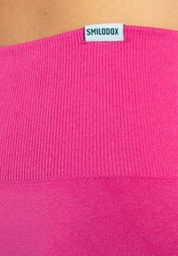 Fuksia urheilu toppi, jossa on ribattu pinta ja sileä alaosa, varustettu sinisellä SMILODOX-logotunnisteella kaula-aukossa.