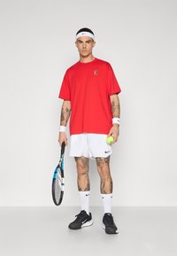 Punainen ylipitkä t-paita, valkoiset shortsit, mustat lenkkarit ja valkoiset tennsockit. Pitelee tennismailaa ja vihreää tennispalloa.
