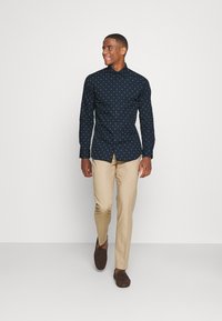 Camicia blu con bottoni, caratterizzata da un motivo a pois bianchi ripetuto, abbinata a pantaloni beige chiaro e mocassini marroni. Il modello sta camminando.