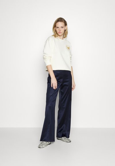 Hanorac alb cu un logo auriu, asortat cu pantaloni largi din satin navy și pantofi slip-on cu model. O ținută simplă și casual.