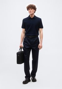 Νέος άνδρας με σγουρά μαλλιά, φορώντας μαύρο πόλο πουκάμισο, μαύρο παντελόνι, μαύρα παπούτσια και κρατώντας μια μαύρη χαρτοφύλακα στο δεξί του χέρι.