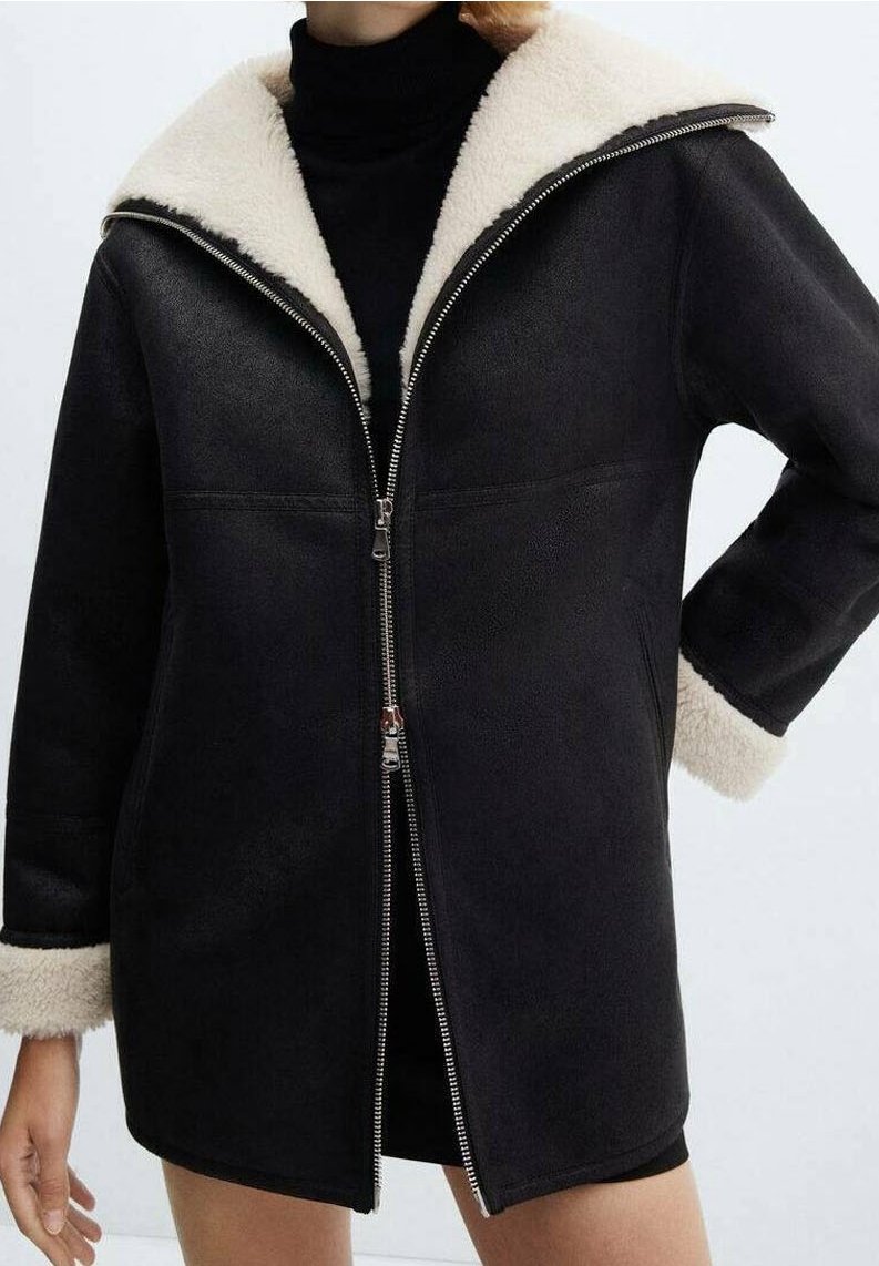 Manteau noir mi-long avec doublure en shearling crème au col et aux poignets, porté ouvert sur une robe noire à col roulé.