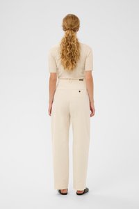 Kvinde med langt bølget hår i en hestehale iført en beige kortærmet bluse, højtaljede cremede bukser og sorte sko, stående med ryggen til.