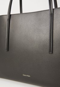 Elegant svart Calvin Klein tote-veske med strukturert lærfinish, utstyrt med to holdbare håndtak og en diskret gravert merkevarelogo.