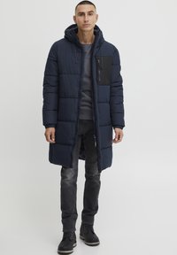 Abrigo acolchado azul marino con capucha, que cuenta con un bolsillo en el pecho con cremallera negra, mangas largas y un diseño hasta el muslo. Llevado con jeans oscuros y botas.