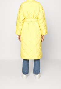Gepolsterte gelbe Oberbekleidung mit einem taillierten Gürtel, weiten Ärmeln und einem knielangen Design. Verfügt über diagonale Nähte und eine glatte Textur.