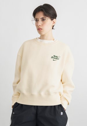 Jaunas asmuo su trumpais tamsiais plaukais, dėvintis skaidraus rėmo akinius, kreminę Nike džemperį su „Nike Summer“ logotipu ir juodus šortus su logotipais.