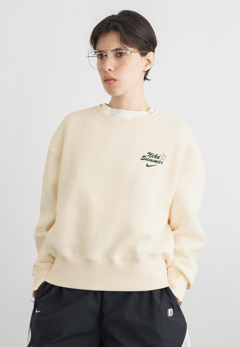 Νεαρό άτομο με κοντά σκούρα μαλλιά, που φοράει γυαλιά με διάφανο πλαίσιο, κρεμ φούτερ Nike με το λογότυπο "Nike Summer" και μαύρο σορτς με λογότυπα.