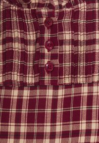 Tissu à carreaux prune et crème avec des lignes verticales et horizontales. Présente une patte de boutonnage avec quatre boutons bordeaux et une texture froncée.