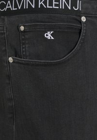 Černé džínové kalhoty mají na zadní kapse malé vyšívané logo "ck" a stříbrné akcenty kolem okrajů kapes.