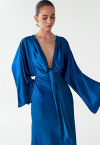 Blauwe satijnen jurk met een diepe V-hals, brede mouwen en een detail van een gestrikte taille. Soepele textuur, vloeiend ontwerp en levendige kleur.