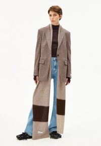 Blazer en mélange de laine marron par-dessus un pull côtelé bordeaux, associé à un jean flare bleu clair et une longue écharpe rayée multicolore.