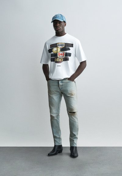 Άνδρας με λευκό γραφικό μπλουζάκι, ανοιχτόχρωμο τζιν με σκισίματα, μαύρα μποτάκια και μπλε καπέλο μπέιζμπολ, στέκεται με τα χέρια στις τσέπες.