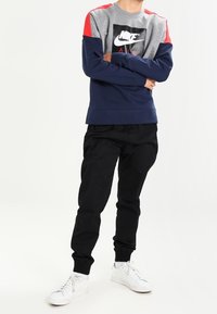 Nike-tröjan har en grå kropp, röda axelklamrar och en marinblå nederdel. Svarta byxor och vita sneakers fulländar outfiten.