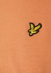 Camiseta de algodón naranja con un logo de pájaro bordado en negro y amarillo en el lado izquierdo. Textura suave con un diseño ajustado.