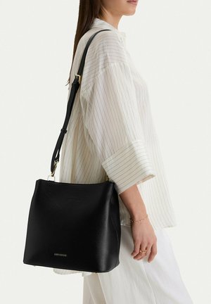 Žena nosící bílou blůzu s proužky a bílé kalhoty, nesoucí černou koženou kabelku s kováním zlaté barvy.