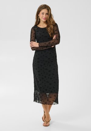 Une femme porte une robe midi noire à fleurs, avec des manches longues et un ourlet transparents. Elle se tient debout, les bras croisés, devant un fond clair et uni.