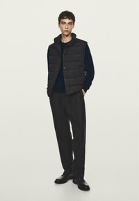 Gilet imbottito nero sopra una maglietta a maniche lunghe blu scuro, abbinato a pantaloni larghi scuri e scarpe nere, su uno sfondo neutro.