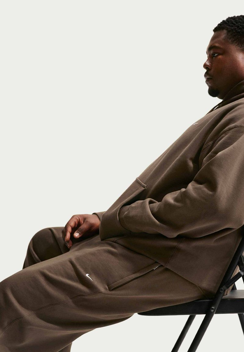 Felpa con cappuccio marrone e pantaloni della tuta in tessuto morbido, con tasca frontale e dettagli laterali, indossati mentre si è seduti su una sedia nera.